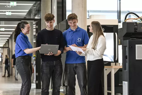 Four young Böllhoff employees are standing together in a modern industrial building discussing something. One person is holding a laptop, another is holding documents. Machines and workstations can be seen in the background, the surroundings appear technical and open.