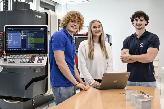 Drei junge Böllhoff Mitarbeitende stehen in einem modernen Technikraum an einem Holztisch. Auf dem Tisch befindet sich ein aufgeklappter Laptop und einige Metallbauteile. Im Hintergrund ist eine große CNC-Maschine mit offenem Bedienfeld und sichtbarem Display zu sehen.