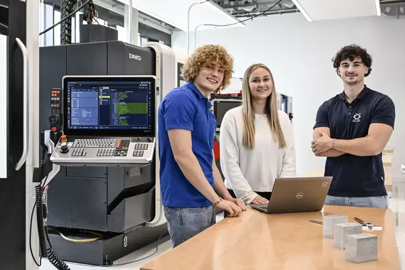 Drei junge Böllhoff Mitarbeitende stehen in einem modernen Technikraum an einem Holztisch. Auf dem Tisch befindet sich ein aufgeklappter Laptop und einige Metallbauteile. Im Hintergrund ist eine große CNC-Maschine mit offenem Bedienfeld und sichtbarem Display zu sehen.