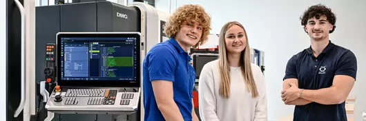 Drei junge Böllhoff Mitarbeitende stehen in einem modernen Technikraum an einem Holztisch. Auf dem Tisch befindet sich ein aufgeklappter Laptop und einige Metallbauteile. Im Hintergrund ist eine große CNC-Maschine mit offenem Bedienfeld und sichtbarem Display zu sehen.