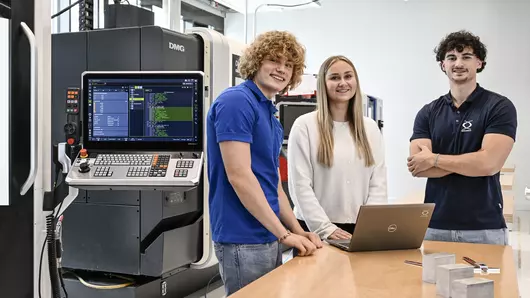 Three young Böllhoff employees are standing at a wooden table in a modern technology room. On the table is an open laptop and some metal components. A large CNC machine with an open control panel and visible display can be seen in the background.
