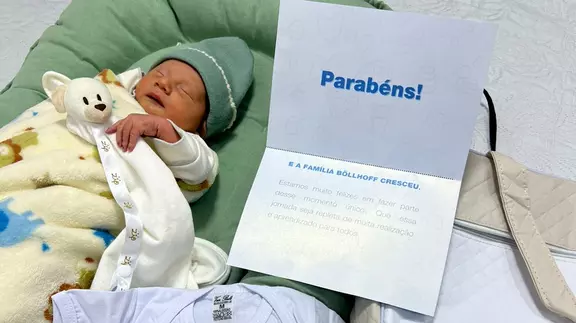 A newborn child with employees of Böllhoff Brazil is wrapped in a blanket and is holding the cuddly toy from the "Kit Maternidade" in its arm. Next to them are the other items from the "Kit Maternidade".
