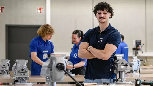 Mit Know-how und Teamgeist: Ein Auszubildender steht in der modernen Werkstatt, während sein Team im Hintergrund praktisch arbeitet.