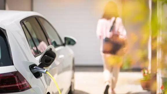 A woman walks away from an electric car in a driveway with the charging plug plugged in.