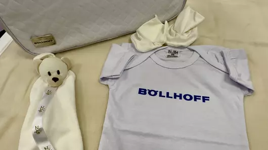 The items in the "Kit Maternidade": a bag, a cuddly toy, a babygro with the Böllhoff logo on the chest, a pair of little socks and a comfort blanket.