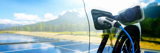 An image of an electric car with a charging plug in front of a green landscape with a photovoltaic system.