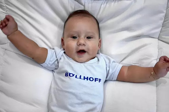 Ein Baby von Mitarbeitern aus Brasilien trägt einen weißen Body mit Böllhoff Logo und schaut gespannt in die Kamera