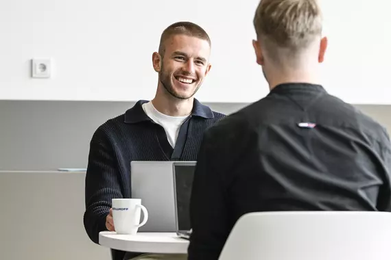 Two young Böllhoff employees sit at a round table in a modern, bright room. In front of one of them is an open laptop and a coffee cup bearing the Böllhoff logo. The atmosphere is calm and focused, as though they were in the middle of a conversation or meeting.