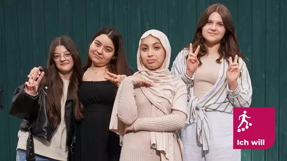Four girls stand in front of a green wooden wall and smile into the camera. The logo of the ‘Ich will’ foundation is at the bottom right of the picture