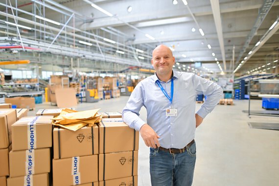 Eine Böllhoff Mitarbeiter steht an Kartons gelehnt im Warenausgang lächelt in die Kamera