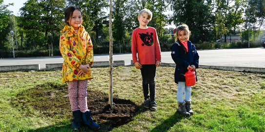 Drei Mitarbeiterkinder stehen neben einem selbst gepflanzten Baum auf dem Böllhoff Werksgelände in Bielefeld.
